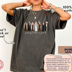 May include: A charcoal gray t-shirt featuring a graphic of seven religious figures in various habits and robes, with the text "pray like a girl" written in a cursive script below the figures. The shirt is made of 100% US cotton and is size XL.