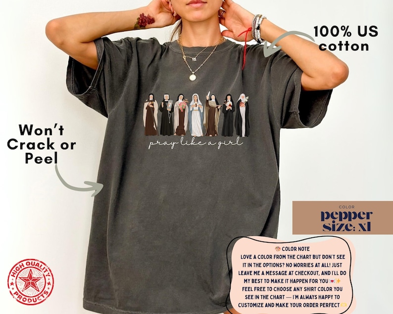 May include: A charcoal gray t-shirt featuring a graphic of seven religious figures in various habits and robes, with the text "pray like a girl" written in a cursive script below the figures. The shirt is made of 100% US cotton and is size XL.