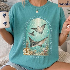May include: A seafoam green t-shirt featuring a graphic of two stingrays swimming underwater. The text reads, "WHEN YOU GO THROUGH DEEP WATERS, I WILL BE WITH YOU" and "ISAIAH 43:2". The design includes coral, bubbles, and an open book with a cross.