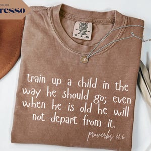 May include: Espresso-colored t-shirt with the text "train up a child in the way he should go; even when he is old he will not depart from it. proverbs 22.6" in white. A silver necklace with a pendant rests on the shirt. The shirt is folded.
