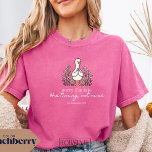 Op de afbeelding: Een roze t-shirt met de tekst "sorry I'm late His timing, not mine Ecclesiastes 3:1" en een cartoon eend omringd door bloemen. Het shirt is effen van kleur en heeft korte mouwen.
