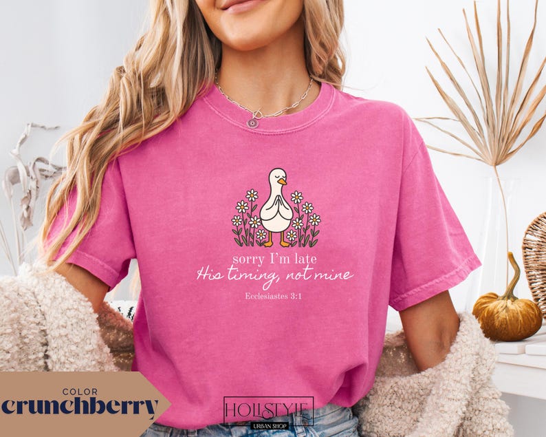Op de afbeelding: Een roze t-shirt met de tekst "sorry I'm late His timing, not mine Ecclesiastes 3:1" en een cartoon eend omringd door bloemen. Het shirt is effen van kleur en heeft korte mouwen.