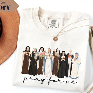 May include: Ivory-colored t-shirt featuring a graphic of several religious figures in various robes and habits. The phrase "pray for us" is written in a cursive font at the bottom of the design. A gold necklace is visible.