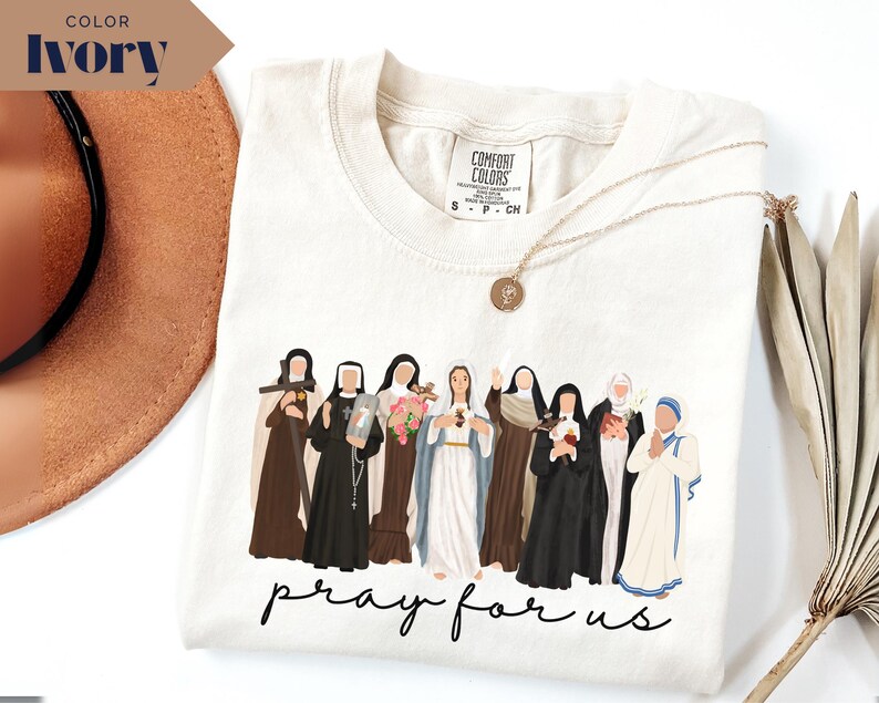 May include: Ivory-colored t-shirt featuring a graphic of several religious figures in various robes and habits. The phrase "pray for us" is written in a cursive font at the bottom of the design. A gold necklace is visible.
