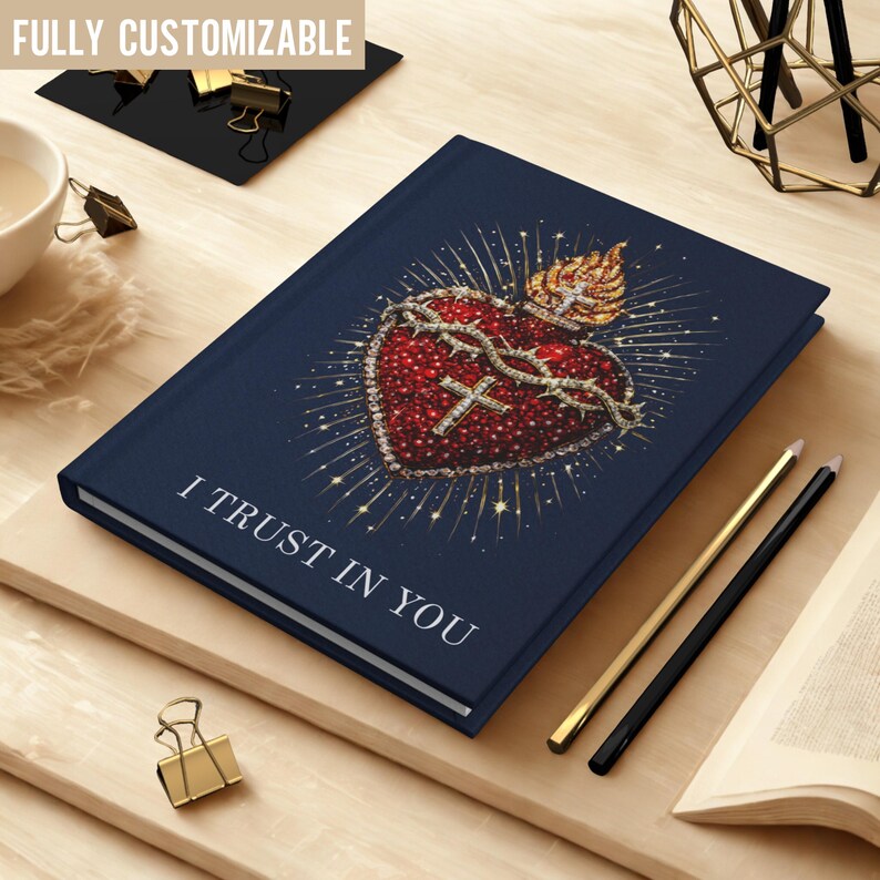 May include: A navy blue journal with the text "I TRUST IN YOU" and a detailed illustration of a Sacred Heart. The heart is red with a crown of thorns and a cross, surrounded by sparkling accents. Gold and black pencils are next to the journal.