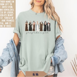 May include: A sage green crewneck t-shirt featuring a graphic of eight religious figures in traditional habits and the text "pray like a girl" printed in black script. The figures are arranged in a row, each holding a distinct religious item.