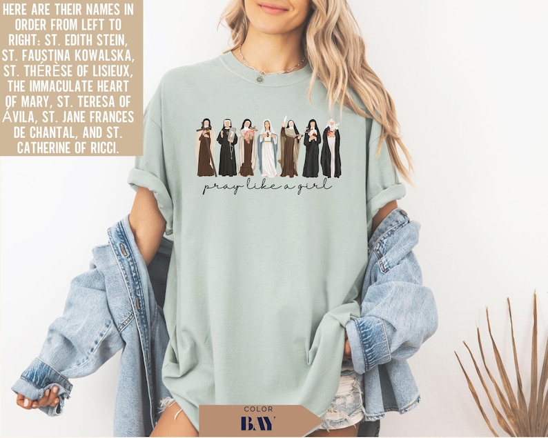 May include: A sage green crewneck t-shirt featuring a graphic of eight religious figures in traditional habits and the text "pray like a girl" printed in black script. The figures are arranged in a row, each holding a distinct religious item.