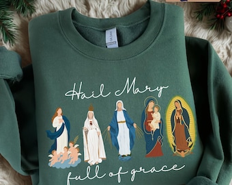 May include: Forest green sweatshirt with the words "Hail Mary full of grace" in white script. The sweatshirt features illustrations of religious figures in various colors. The color of the sweatshirt is labeled "forest green" in a small box in the upper right corner.