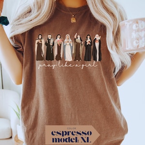 May include: An espresso-colored t-shirt featuring a graphic illustration of seven religious figures in traditional habits and robes. Below the figures, the text "pray like a girl" is printed in a white cursive script. The shirt has a crew neckline.
