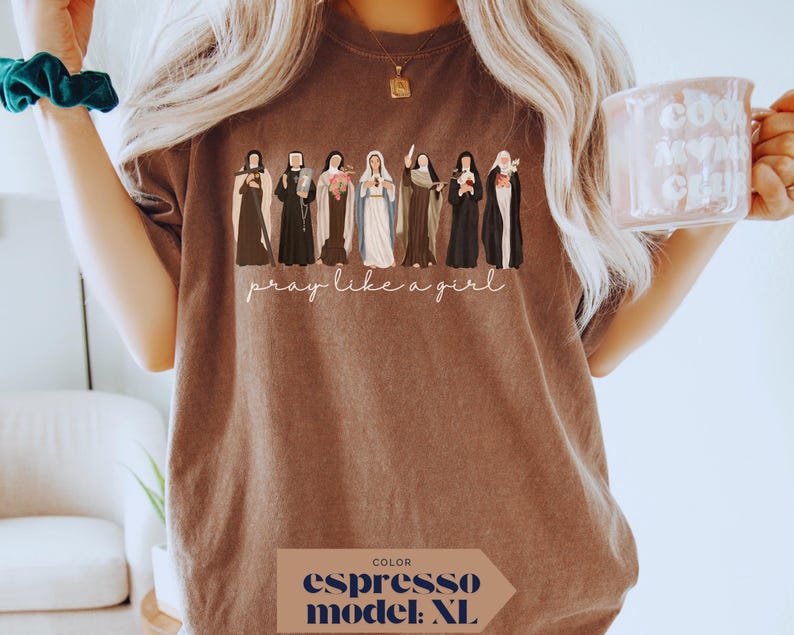 May include: An espresso-colored t-shirt featuring a graphic illustration of seven religious figures in traditional habits and robes. Below the figures, the text "pray like a girl" is printed in a white cursive script. The shirt has a crew neckline.