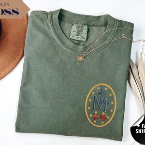 Pu&ograve; includere: Una maglietta verde muschio con una toppa ovale ricamata con una croce, le lettere "M" e "F" e due cuori. Una collana d'oro poggia sulla maglietta. Le parole "COLOR MOSS" sono in uno striscione.