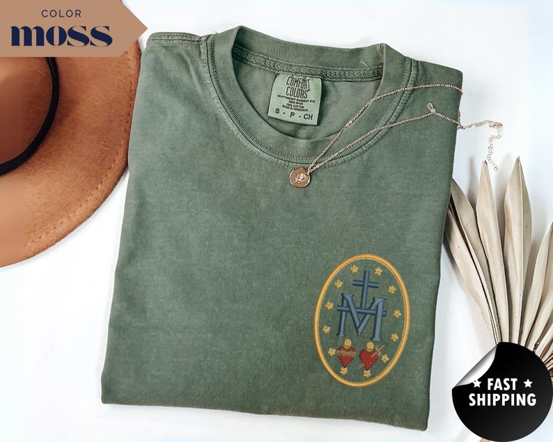 Pu&ograve; includere: Una maglietta verde muschio con una toppa ovale ricamata con una croce, le lettere "M" e "F" e due cuori. Una collana d'oro poggia sulla maglietta. Le parole "COLOR MOSS" sono in uno striscione.