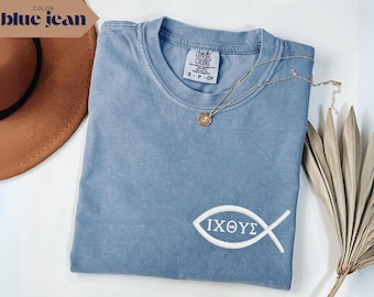 May include: A blue jean colored t-shirt with a white embroidered Ichthys fish symbol. The shirt has a crew neck and short sleeves. A gold necklace with a pendant and a brown hat are also in the image.