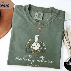 Op de afbeelding: Een mosgroen Comfort Colors T-shirt met een cartoon eend ontwerp, omringd door madeliefjes en de tekst "sorry I'm late, His timing, not mine, Ecclesiastes 3:1". Een gouden ketting ligt op het shirt.