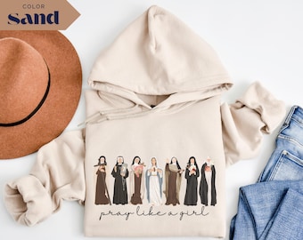 Puede incluir: Sudadera con capucha color arena con cordón y un gráfico de varias mujeres con vestimenta religiosa. La frase "pray like a girl" está impresa en una fuente de escritura debajo del gráfico. También se ven un sombrero marrón y unos vaqueros azules.