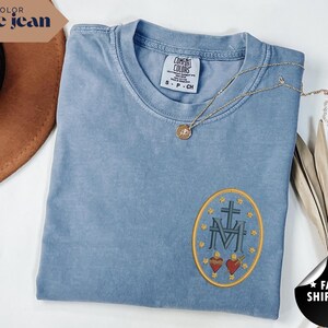 Pu&ograve; includere: Una maglietta Comfort Colors blu jeans con un design ovale ricamato con una croce, le lettere "M" e "V" e due cuori. Una collana d'oro con un ciondolo poggia sulla maglietta. La maglietta &egrave; piegata e viene esposta con un cappello marrone e una pianta secca.
