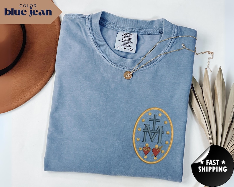 Pu&ograve; includere: Una maglietta Comfort Colors blu jeans con un design ovale ricamato con una croce, le lettere "M" e "V" e due cuori. Una collana d'oro con un ciondolo poggia sulla maglietta. La maglietta &egrave; piegata e viene esposta con un cappello marrone e una pianta secca.