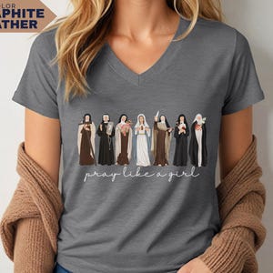 Op de afbeelding: Grafietkleurig V-hals T-shirt met een illustratie van religieuze figuren. De tekst "pray like a girl" staat in cursief onder de figuren. Het shirt heeft een neutrale grijze kleur.