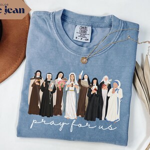 May include: A blue jean colored t-shirt featuring a graphic of several religious figures. The phrase "pray for us" is printed in white script at the bottom. A gold necklace with a pendant rests on the shirt.