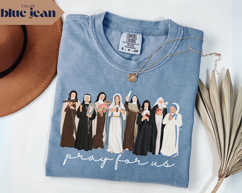 May include: A blue jean colored t-shirt featuring a graphic of several religious figures. The phrase "pray for us" is printed in white script at the bottom. A gold necklace with a pendant rests on the shirt.