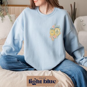May include: Light blue sweatshirt with a floral and religious emblem design. The emblem features a gold-framed image and a circular design with the letters "M" and "A". The sweatshirt is a light blue color, with the text "light blue" at the bottom.