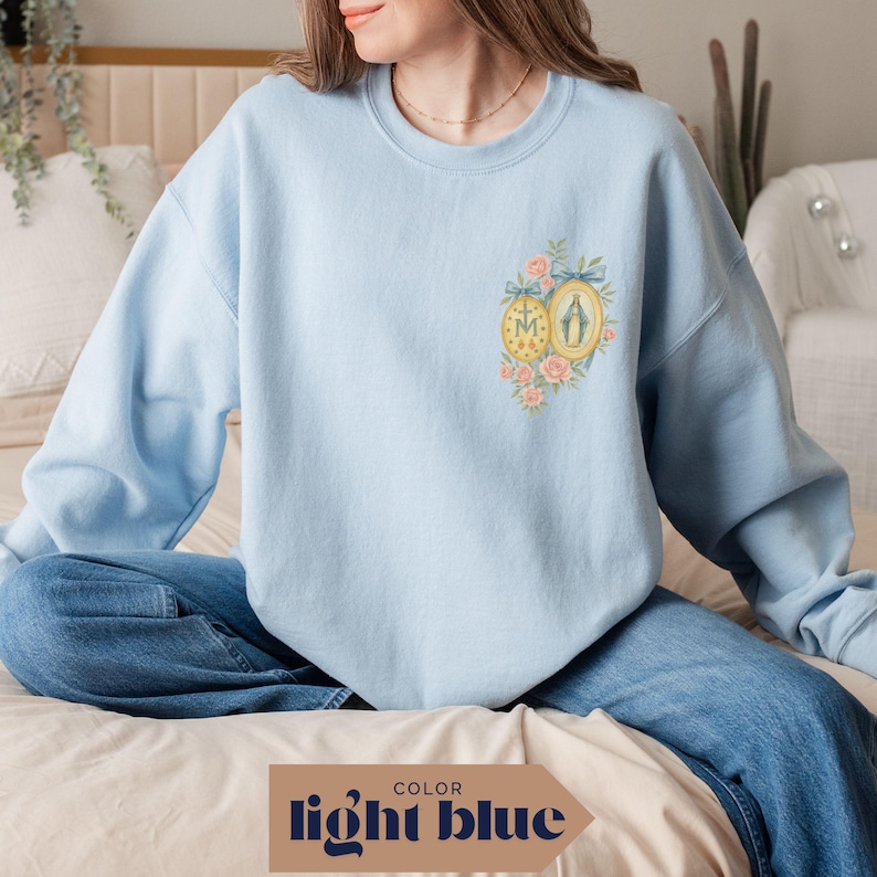 May include: Light blue sweatshirt with a floral and religious emblem design. The emblem features a gold-framed image and a circular design with the letters "M" and "A". The sweatshirt is a light blue color, with the text "light blue" at the bottom.