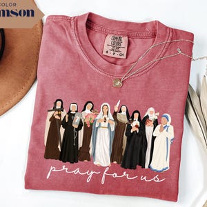 May include: Crimson-colored t-shirt featuring an illustration of several religious figures. The phrase "pray for us" is printed in white script below the figures. A gold necklace with a pendant rests on the shirt. The shirt is a Comfort Colors brand.