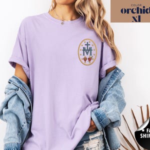 Pu&ograve; includere: Una maglietta lilla chiaro con maniche arrotolate. La maglietta presenta un disegno ricamato di una croce, la lettera M e due cuori all'interno di una cornice ovale. Il testo "COLOR orchid XL" si trova nell'angolo in alto a destra.