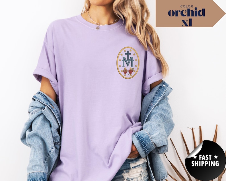 Pu&ograve; includere: Una maglietta lilla chiaro con maniche arrotolate. La maglietta presenta un disegno ricamato di una croce, la lettera M e due cuori all'interno di una cornice ovale. Il testo "COLOR orchid XL" si trova nell'angolo in alto a destra.