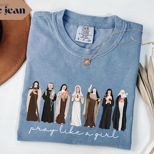 May include: A blue jean colored t-shirt featuring a graphic of seven nuns in various habits with the text "pray like a girl" in white script. The shirt has a crew neckline and a Comfort Colors brand label visible at the inner collar area.