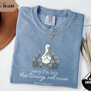 Op de afbeelding: Blauwe jeanskleurige t-shirt met een afbeelding van een witte eend met gevouwen handen, omringd door madeliefjes. De tekst "sorry I'm late, His timing, not mine" staat eronder, met "Ecclesiastes 3:1". Een gouden ketting en een bruine hoed zijn ook zichtbaar.