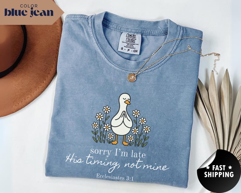 Op de afbeelding: Blauwe jeanskleurige t-shirt met een afbeelding van een witte eend met gevouwen handen, omringd door madeliefjes. De tekst "sorry I'm late, His timing, not mine" staat eronder, met "Ecclesiastes 3:1". Een gouden ketting en een bruine hoed zijn ook zichtbaar.