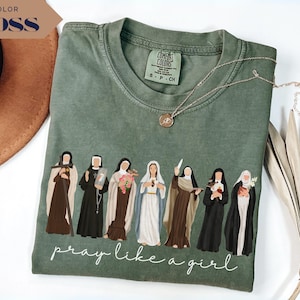 May include: A moss green t-shirt featuring a graphic of seven religious figures in various habits and robes. Below the figures, the text reads "pray like a girl" in a white cursive script. The shirt has a crew neckline and a Comfort Colors brand label.