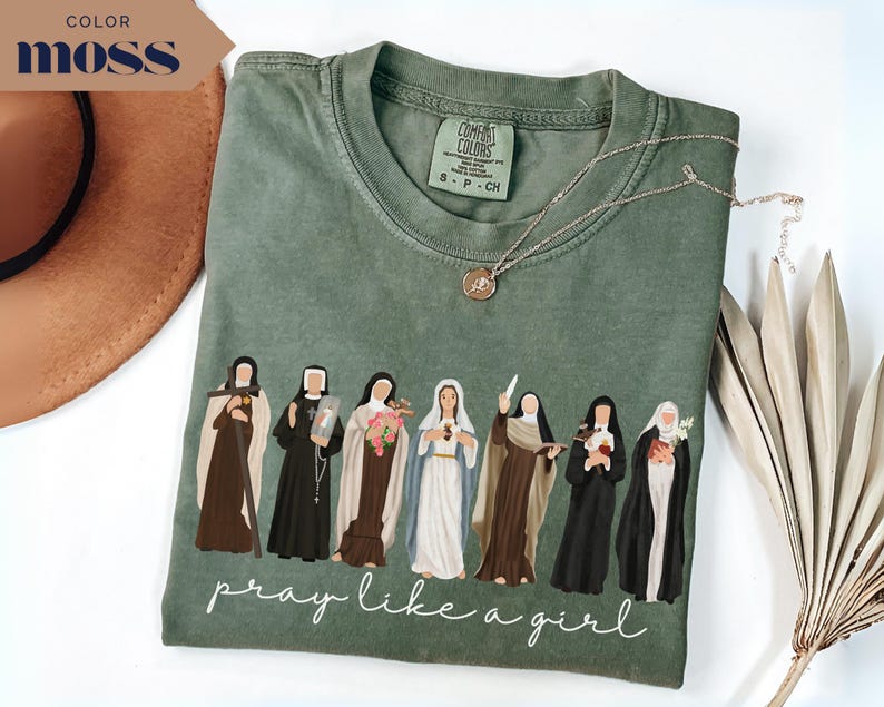 May include: A moss green t-shirt featuring a graphic of seven religious figures in various habits and robes. Below the figures, the text reads "pray like a girl" in a white cursive script. The shirt has a crew neckline and a Comfort Colors brand label.