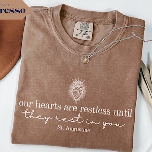 May include: Espresso-colored t-shirt with the text "our hearts are restless until they rest in you" and a heart illustration. A silver necklace with a pendant rests on the shirt. The shirt is a Comfort Colors brand.