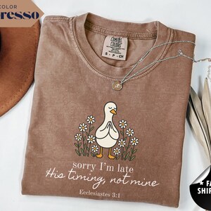 Op de afbeelding: Espresso-kleurige t-shirt met een afbeelding van een witte eend met gevouwen handen, omringd door madeliefjes. De tekst "sorry I'm late, His timing, not mine, Ecclesiastes 3:1" staat eronder. Een zilveren ketting ligt over het shirt.