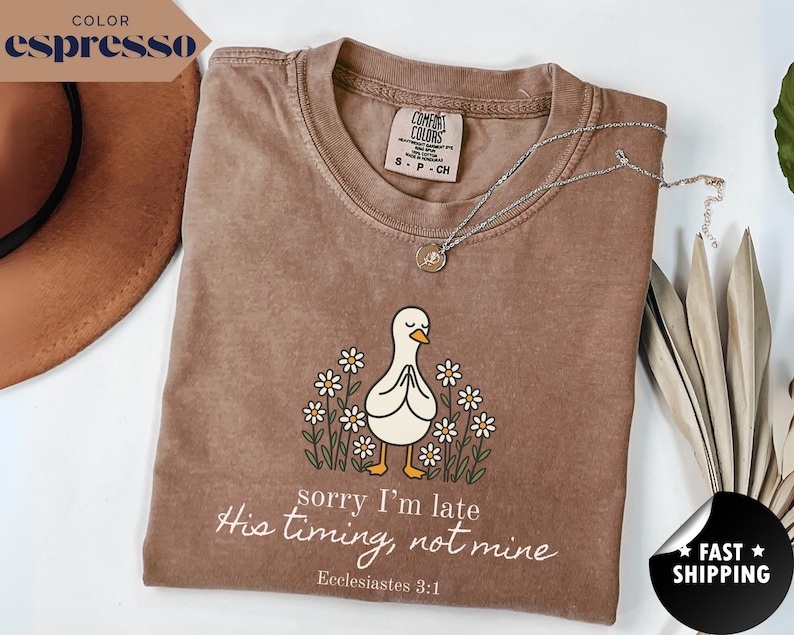 Op de afbeelding: Espresso-kleurige t-shirt met een afbeelding van een witte eend met gevouwen handen, omringd door madeliefjes. De tekst "sorry I'm late, His timing, not mine, Ecclesiastes 3:1" staat eronder. Een zilveren ketting ligt over het shirt.
