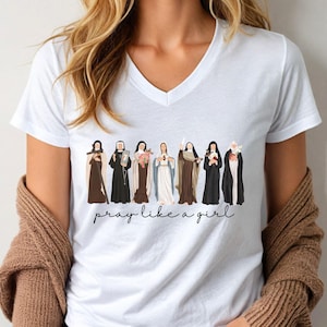 Op de afbeelding: Wit V-hals T-shirt met een afbeelding van verschillende figuren in religieuze kleding. De figuren zijn in verschillende kleuren en de tekst "pray like a girl" staat eronder in cursief. Het T-shirt wordt door een persoon gedragen.