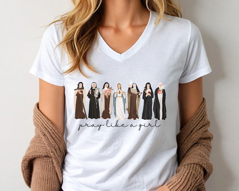 Op de afbeelding: Wit V-hals T-shirt met een afbeelding van verschillende figuren in religieuze kleding. De figuren zijn in verschillende kleuren en de tekst "pray like a girl" staat eronder in cursief. Het T-shirt wordt door een persoon gedragen.