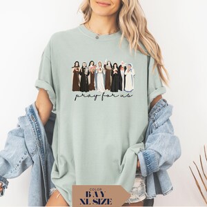 May include: A light green t-shirt featuring a graphic of several figures in various robes. The text "pray for us" is written in a cursive font below the graphic. The shirt is a relaxed fit.