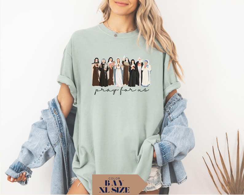 May include: A light green t-shirt featuring a graphic of several figures in various robes. The text "pray for us" is written in a cursive font below the graphic. The shirt is a relaxed fit.