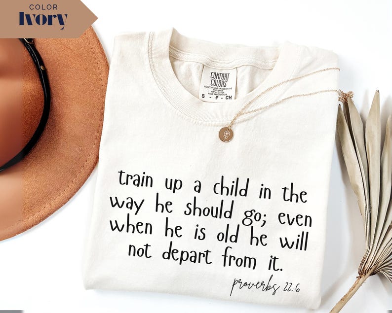May include: Ivory-colored t-shirt with the text "train up a child in the way he should go; even when he is old he will not depart from it. Proverbs 22.6." A gold necklace is draped on the shirt.