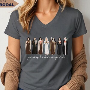 Op de afbeelding: Antracietgrijs V-hals T-shirt met een illustratie van verschillende religieuze figuren. De zin "pray like a girl" staat eronder in cursief. Het woord "CHARCOAL" staat in een kader linksboven.
