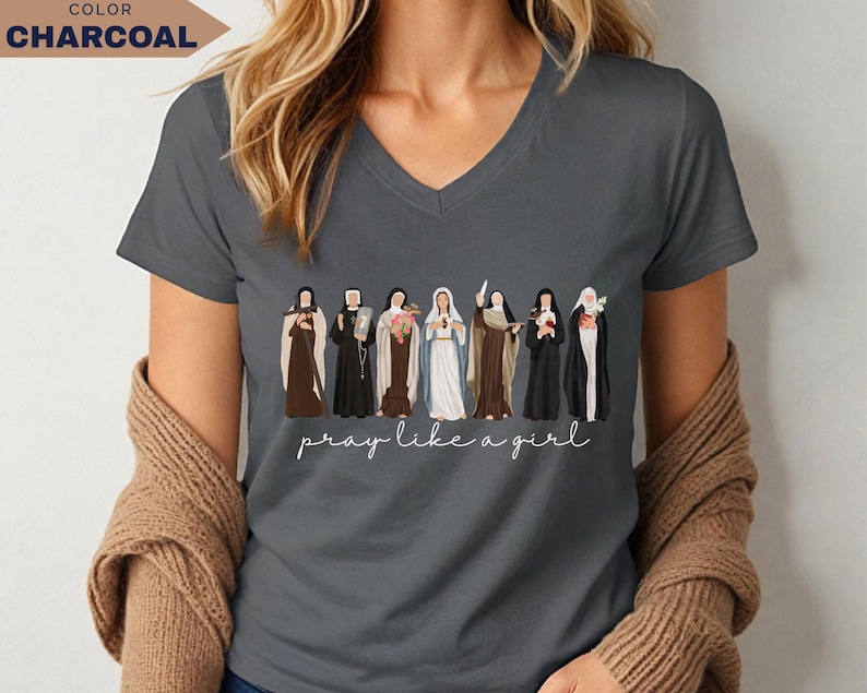 Op de afbeelding: Antracietgrijs V-hals T-shirt met een illustratie van verschillende religieuze figuren. De zin "pray like a girl" staat eronder in cursief. Het woord "CHARCOAL" staat in een kader linksboven.