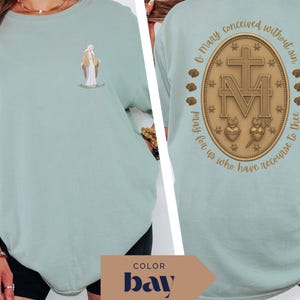May include: A light green t-shirt featuring a small illustration of a religious figure on the front. The back of the shirt displays a gold-colored emblem with a cross, the letter "M", and the text "O Mary conceived without sin, pray for us who have recourse to thee."