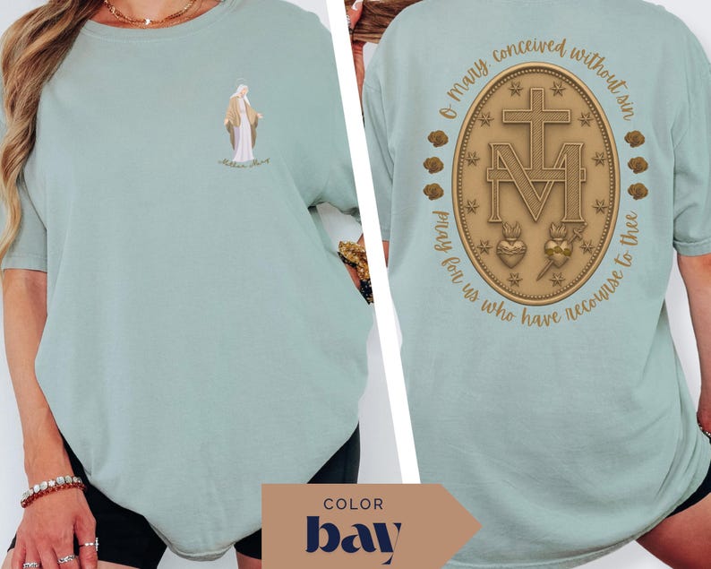 May include: A light green t-shirt featuring a small illustration of a religious figure on the front. The back of the shirt displays a gold-colored emblem with a cross, the letter "M", and the text "O Mary conceived without sin, pray for us who have recourse to thee."