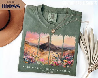 Puede incluir: Camiseta verde musgo con un diseño de paisaje que presenta una cruz en una colina, flores coloridas y el texto "MY SIN WAS GREAT, HIS LOVE WAS GREATER John 3:16". También se ven un collar dorado y un sombrero marrón.
