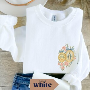 May include: White sweatshirt with a floral and religious design. The design features two oval medallions with a cross and a depiction of the Virgin Mary, surrounded by pink roses and blue ribbons. The sweatshirt is folded on a wooden surface.