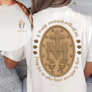 May include: Cream-colored t-shirt featuring a small illustration of a religious figure on the front. The back displays a gold-colored oval emblem with a cross, the letters "M" and "A", and the text "O Mary conceived without sin, pray for us who have recourse to thee."