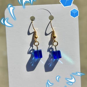 Puede incluir: Un par de pendientes dorados con cuentas cuadradas azules. Los pendientes cuelgan de ganchos dorados y están sujetos a una tarjeta blanca. Las cuentas azules son de un color vibrante. El fondo está borroso.